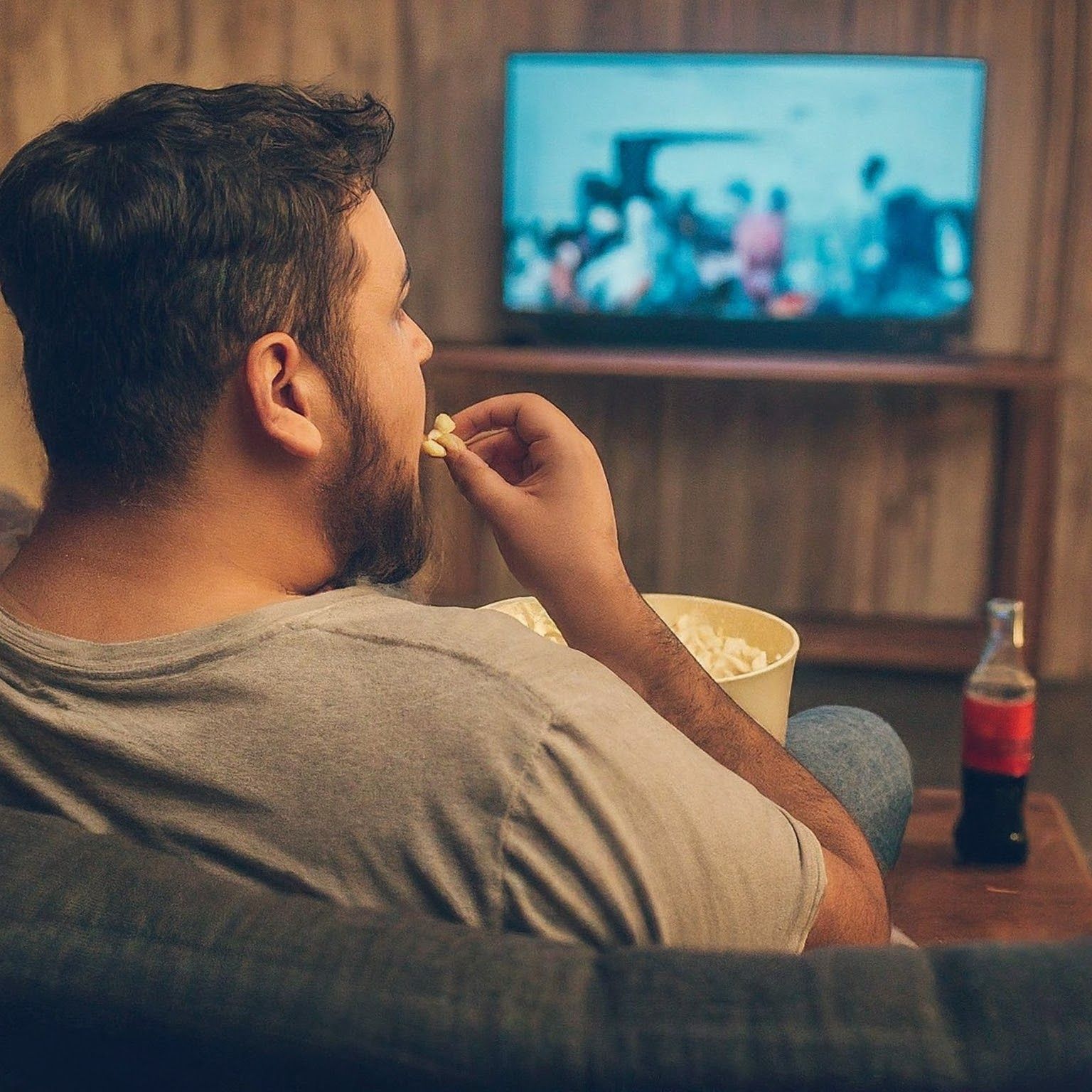 Obese man watching TV and eating popcorn, displaying typical sedentary behavior